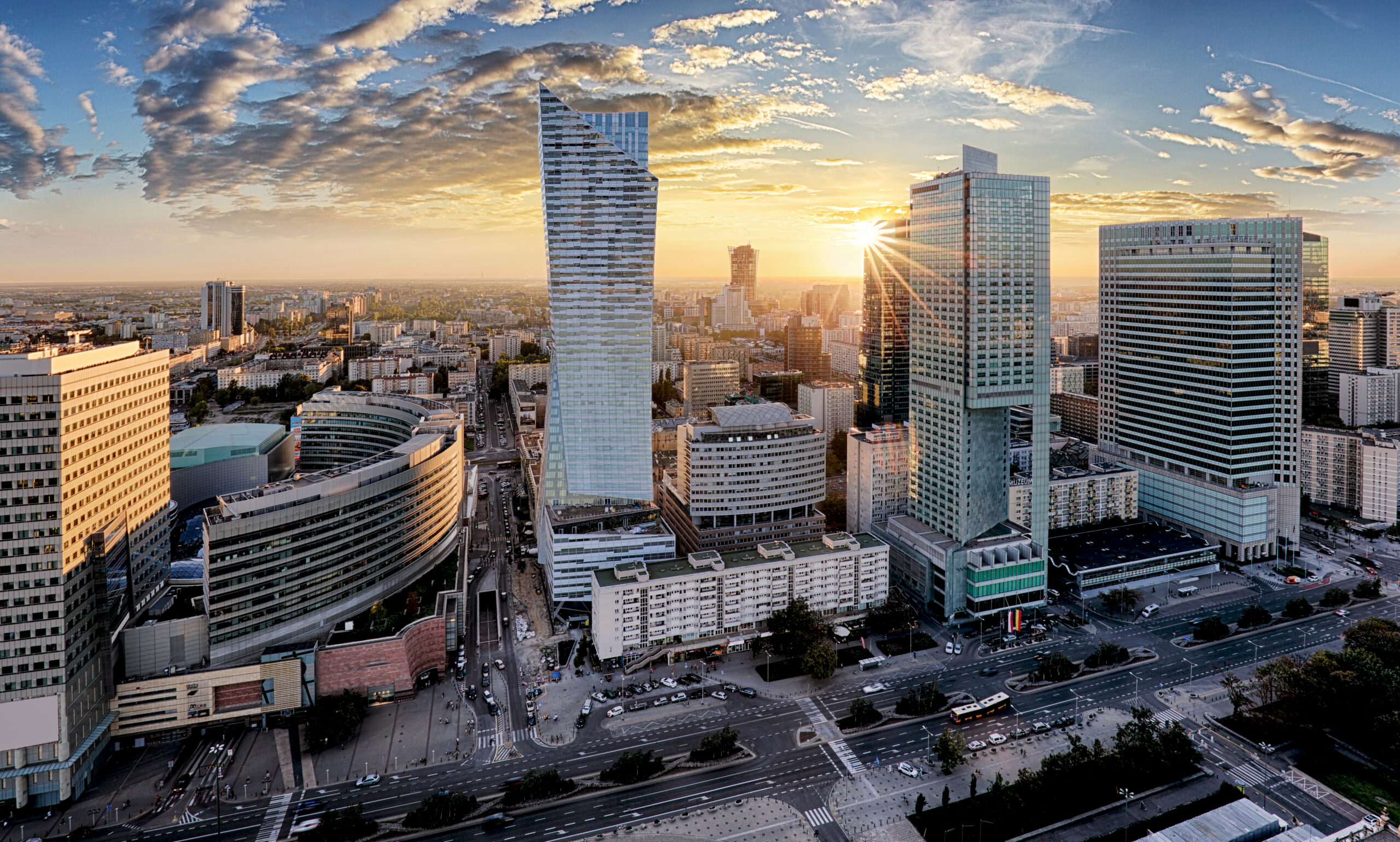 Widok z lotu ptaka na panoramę miasta o zachodzie słońca, z nowoczesnymi wieżowcami ze szklanymi fasadami odbijającymi światło słoneczne. Niebo jest częściowo zachmurzone, rzucając ciepły blask na budynki i ruchliwe ulice. Ukryta poniżej restauracja Konstancin wabi gości swoim czarującym urokiem pośród miejskiej panoramy.