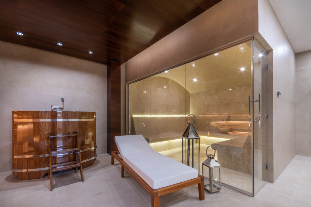 Luksusowy pokój spa z drewnianą sauną, szklanymi drzwiami i eleganckim oświetleniem. Niedaleko wyściełana drewniana leżanka uzupełnia dekoracyjne latarnie, przywołując spokojną atmosferę ekskluzywnej restauracji w Konstancinie. Elegancki drewniany blat ma stalowy kran.