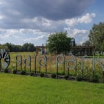 Duży zewnętrzny znak z napisem „gaia park” z logo drzewa stoi pośród trawiastego terenu, z którego wyłania się elegancka sylwetka „luksusowego domu”. W tle pod częściowo zachmurzonym niebem widać nowoczesny budynek i drzewa.