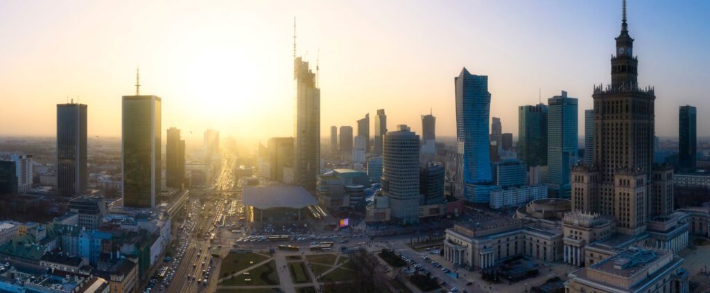Panoramiczny widok na panoramę Warszawy o zachodzie słońca, z wysokimi wieżowcami i budynkami kulturalnymi rzucającymi długie cienie. Zachodzące słońce tworzy ciepłą, jasną atmosferę, rysując sylwetki budynków na tle czystego nieba. Na ulicach poniżej panuje ruch uliczny.