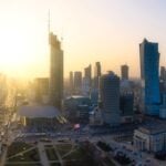 Panoramiczny widok na panoramę Warszawy o zachodzie słońca, z wysokimi wieżowcami i budynkami kulturalnymi rzucającymi długie cienie. Zachodzące słońce tworzy ciepłą, jasną atmosferę, rysując sylwetki budynków na tle czystego nieba. Na ulicach poniżej panuje ruch uliczny.