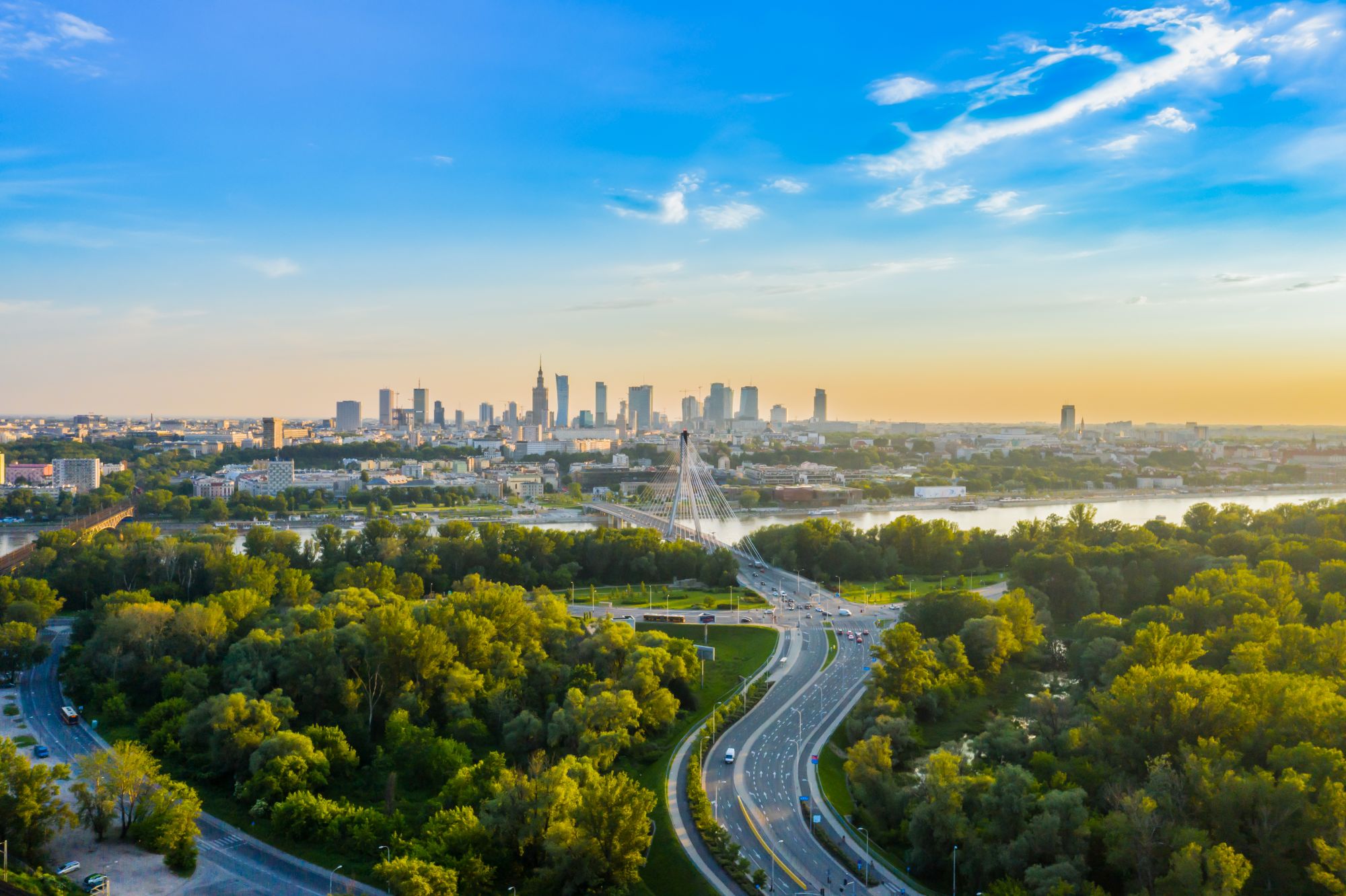 Widok z lotu ptaka na panoramę Warszawy o zachodzie słońca, z rzeką, bujną zielenią drzew, krętymi drogami i licznymi wysokimi budynkami na tle błękitnego nieba z rozproszonymi chmurami.