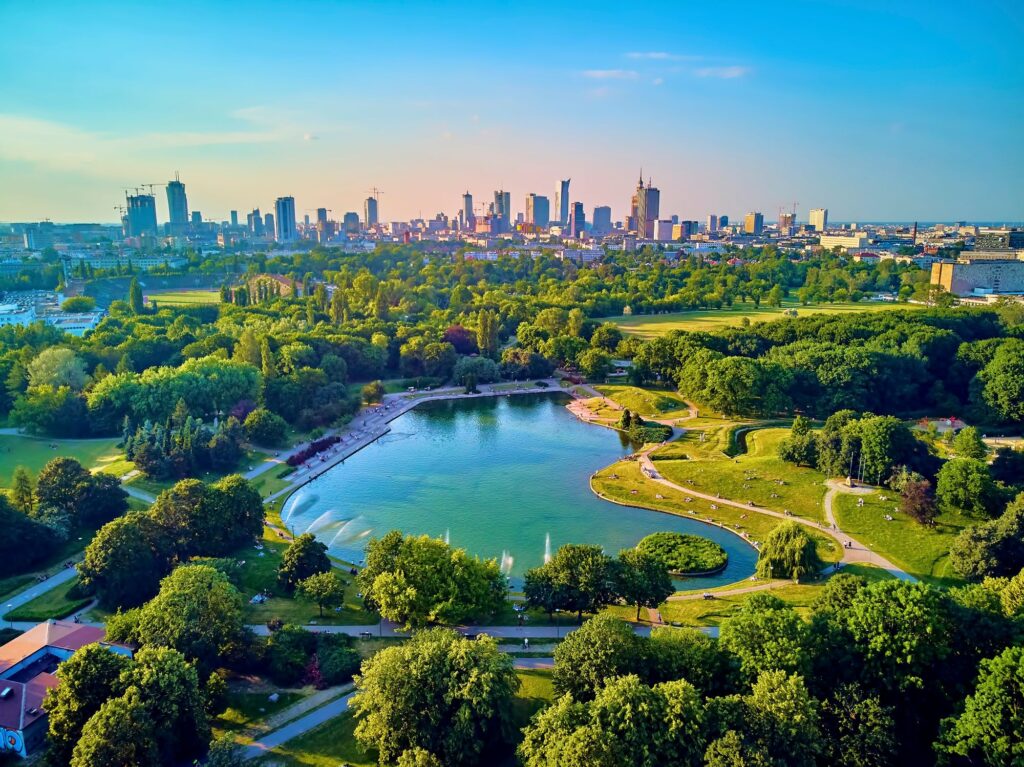 Widok z lotu ptaka na Pola Mokotowskie w Warszawie z jeziorem pośrodku, otoczony drzewami i ścieżkami spacerowymi, na tle panoramy miasta i wysokich budynków pod czystym, błękitnym niebem.