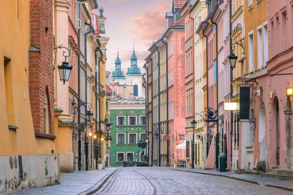 Brukowana ulica, wzdłuż której stoją kolorowe, zabytkowe budynki, prowadzi do zielonego budynku z wieżami i kopułą kościoła w tle pod pastelowym niebem.