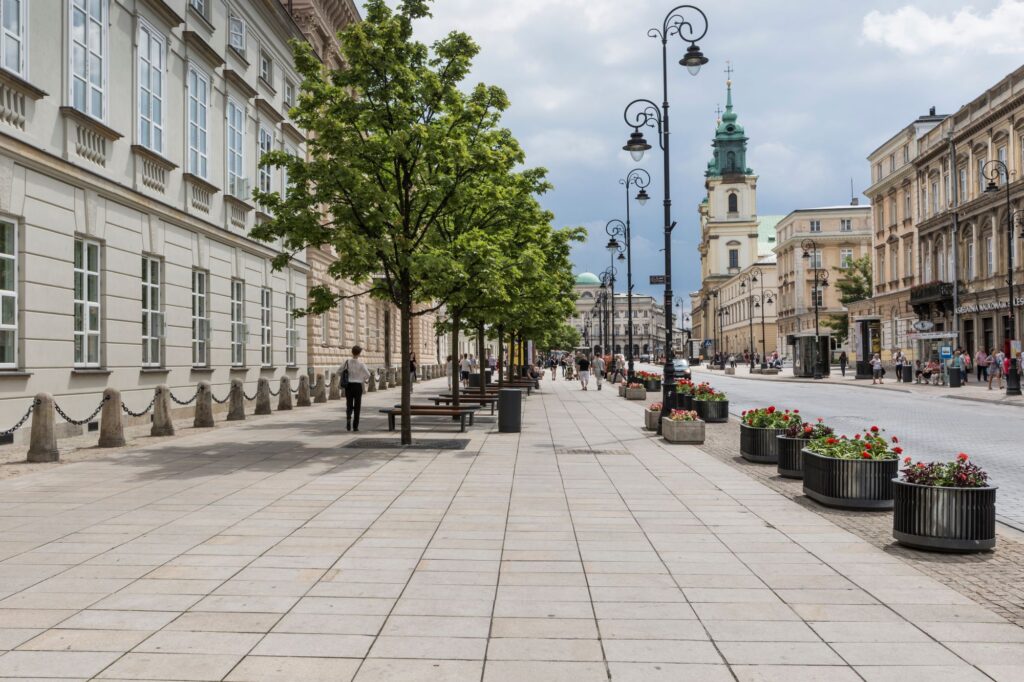 Szeroki, pusty chodnik otoczony drzewami i ławkami biegnie wzdłuż zabytkowych budynków. Dalej na ulicy, pod zachmurzonym niebem, widać kościół z zieloną wieżą.