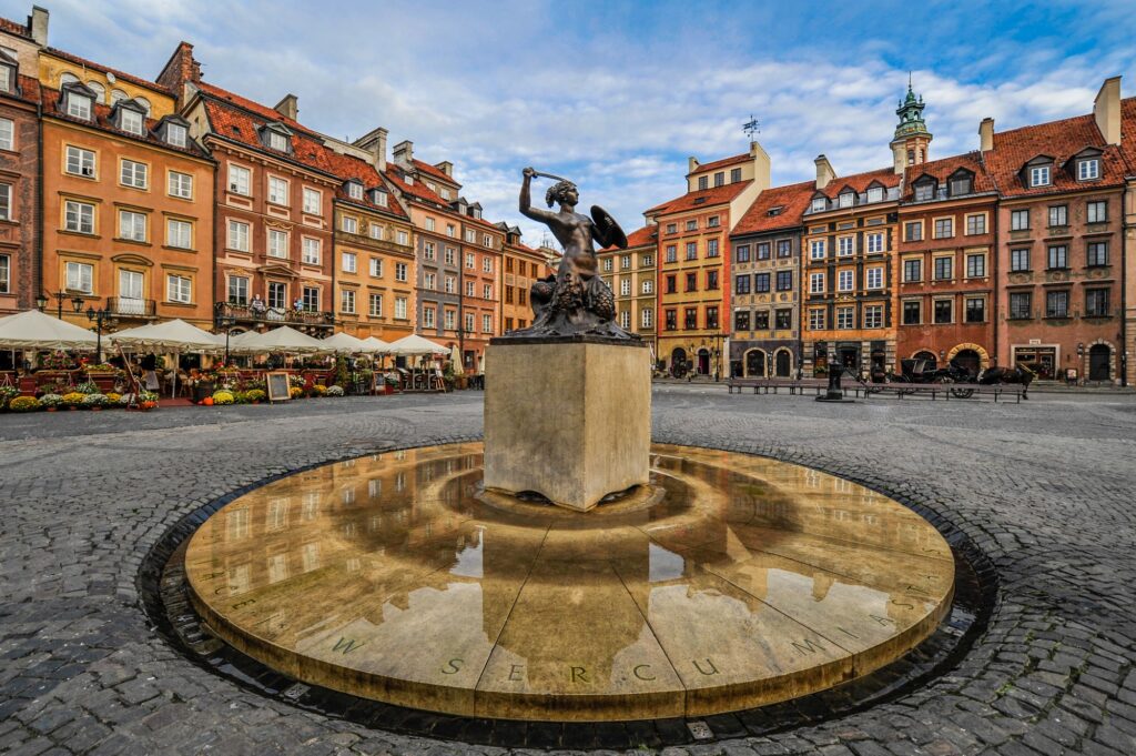 Pośrodku fontanny, na brukowanym placu, stoi posąg syreny z mieczem i tarczą. Wokół placu znajdują się kolorowe, zabytkowe budynki i kawiarnie na świeżym powietrzu, a niebo jest częściowo zachmurzone.