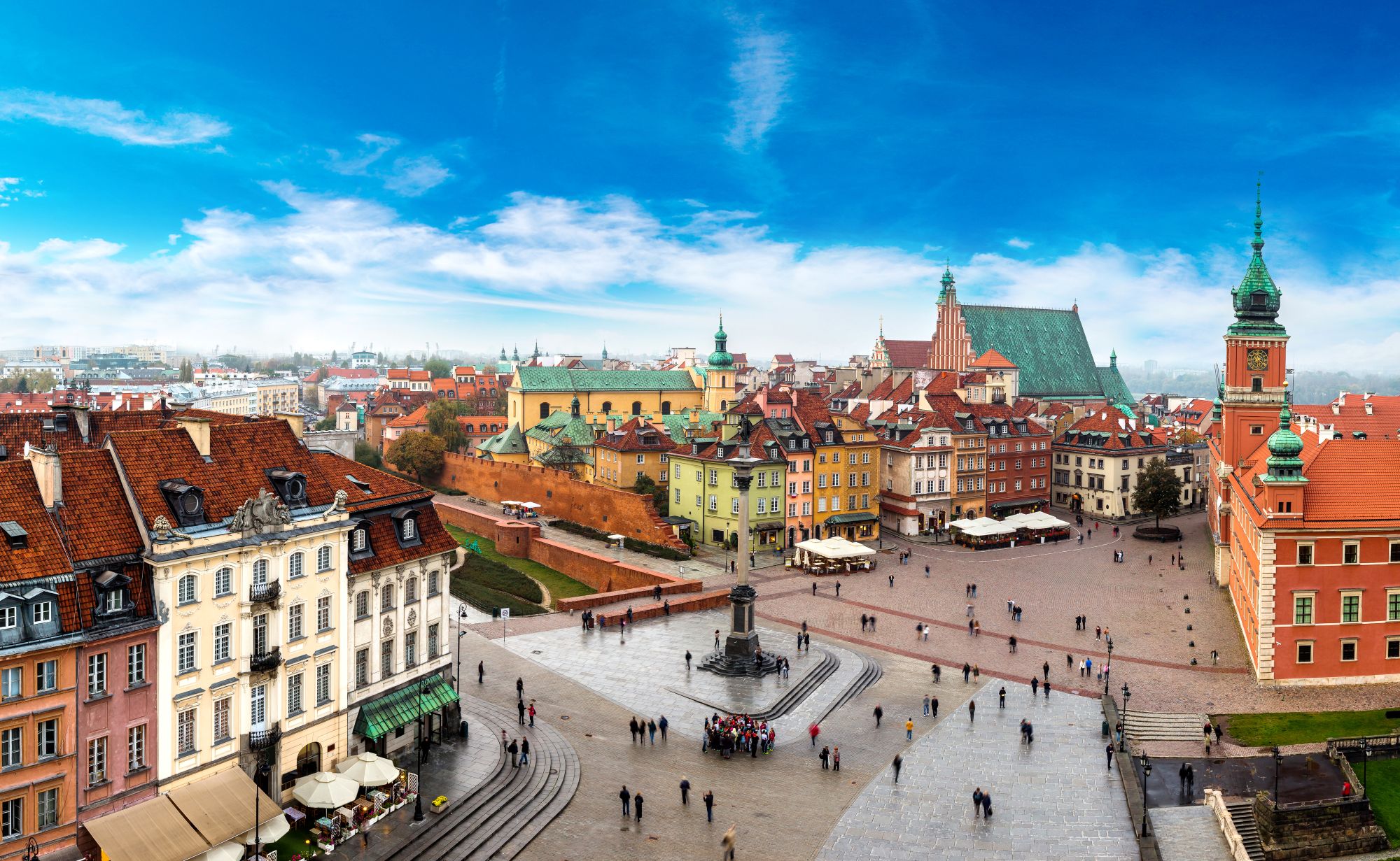 Panoramiczny widok na warszawski Rynek Starego Miasta z kolorowymi, zabytkowymi budynkami, pomnikiem w centrum, spacerującymi ludźmi i dużym kościołem z zielonym dachem pod jasnoniebieskim niebem z rozproszonymi chmurami.