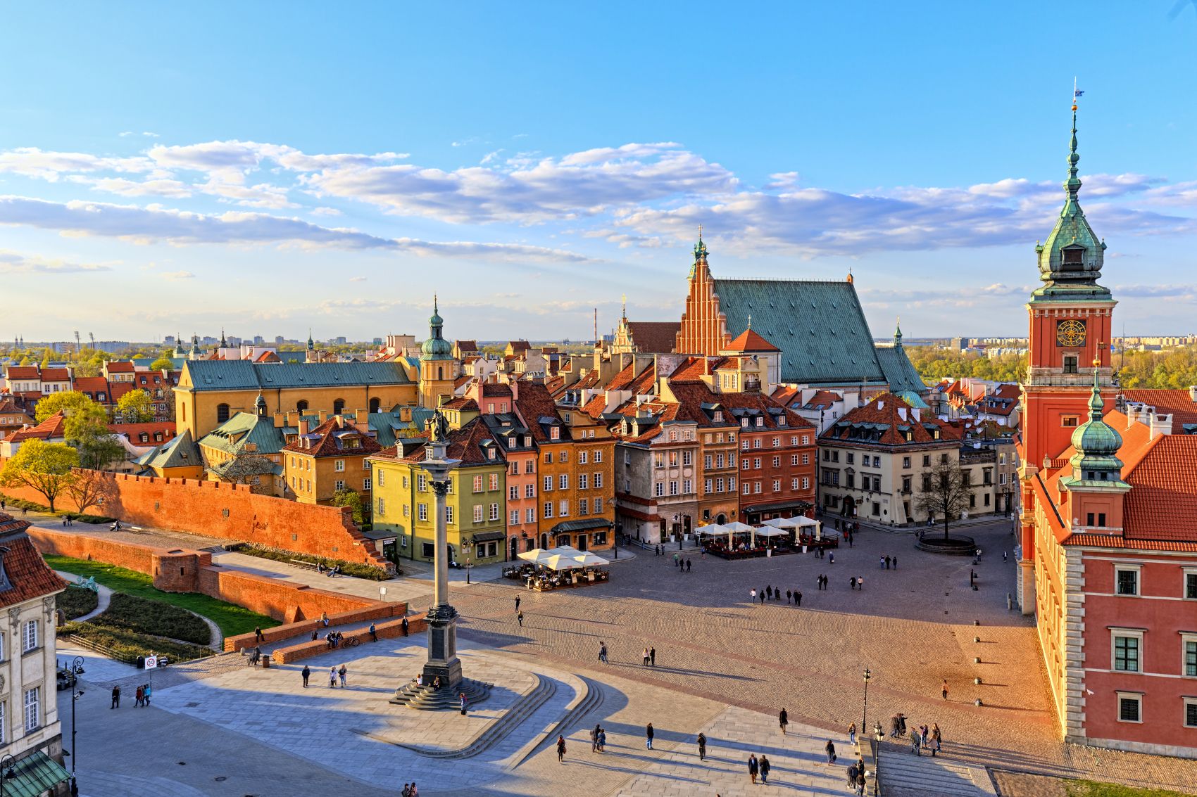 Widok z lotu ptaka na Rynek Starego Miasta w Warszawie z kolorowymi zabytkowymi budynkami, pomnikiem na centralnej kolumnie i dużym kościołem z zielonym dachem na tle błękitnego nieba z rozproszonymi chmurami.