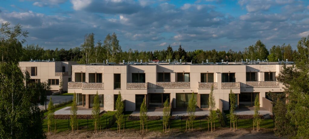 Nowoczesne apartamentowce z płaskimi dachami i balustradami balkonowymi, otoczone drzewami i zielenią pod częściowo zachmurzonym niebem.