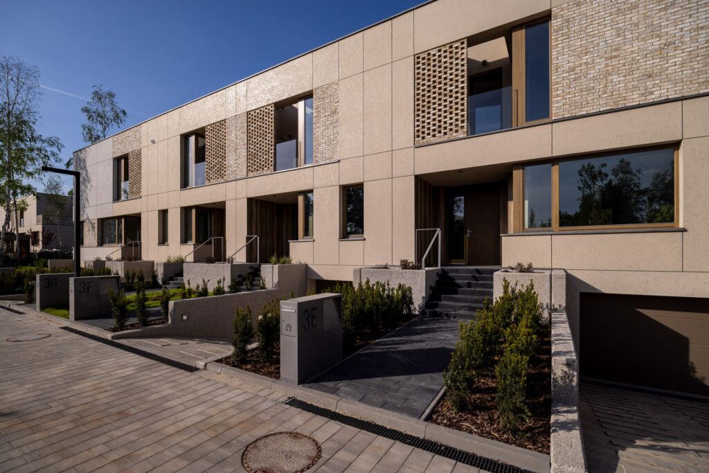 Nowoczesny beżowy kompleks kamienic z geometrycznymi detalami architektonicznymi, dużymi oknami, małymi ogródkami frontowymi i wybrukowanym podjazdem pod czystym błękitnym niebem.