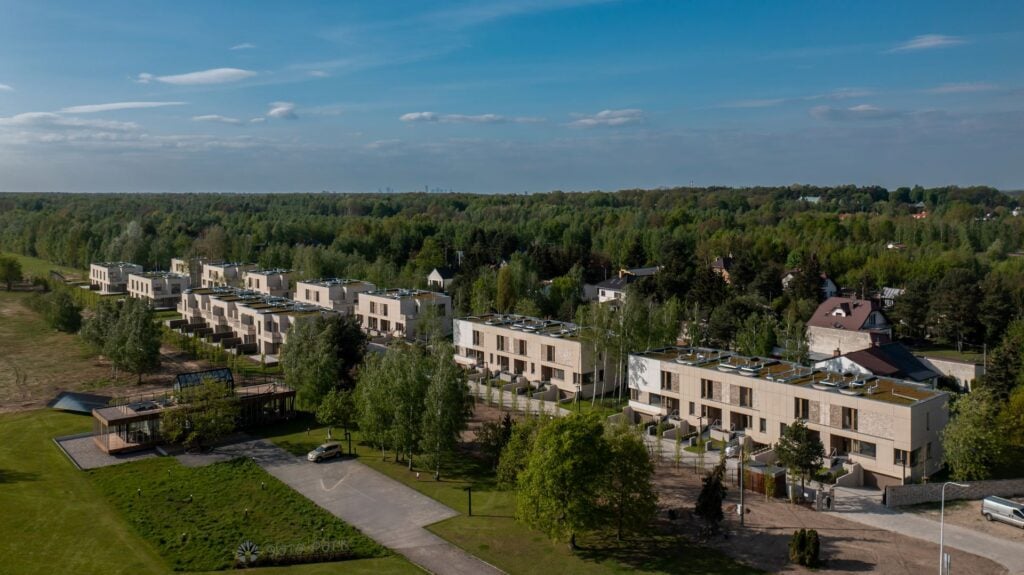 Widok z lotu ptaka na nowoczesne kamienice otoczone zielenią i drzewami, z czystym niebem powyżej i wiejskim krajobrazem w tle.
